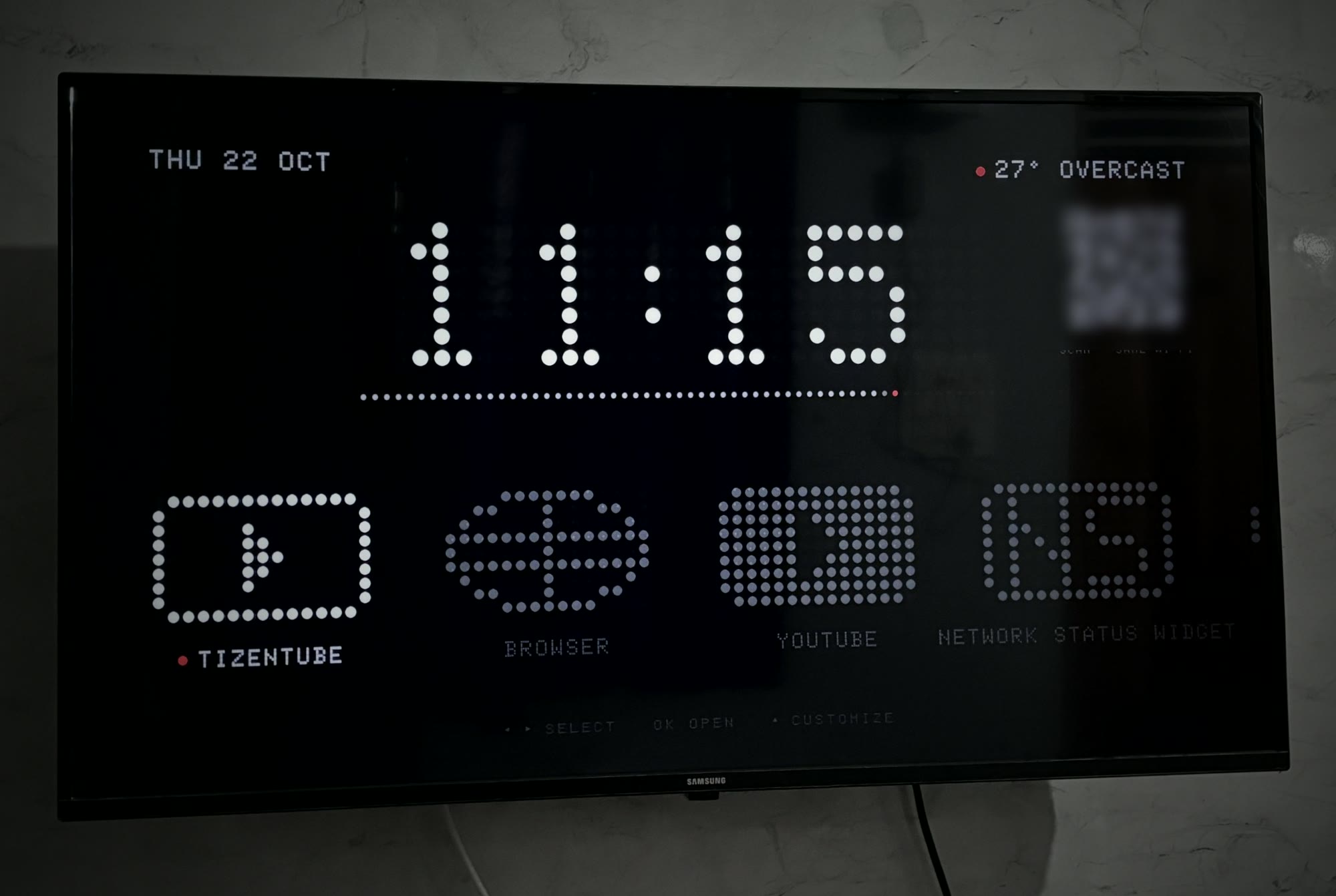 The dot-matrix home screen running on a Samsung TV mounted on a marble wall: date, a large dot clock reading 11:15, weather, and a dock of dot icons.